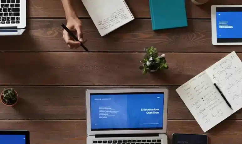 bovenaanzicht van tafel met laptops en mensen die daar aan werken, blaadjes agenda's, telefoons pennen in de hand en andere devices op tafel en in de hand.